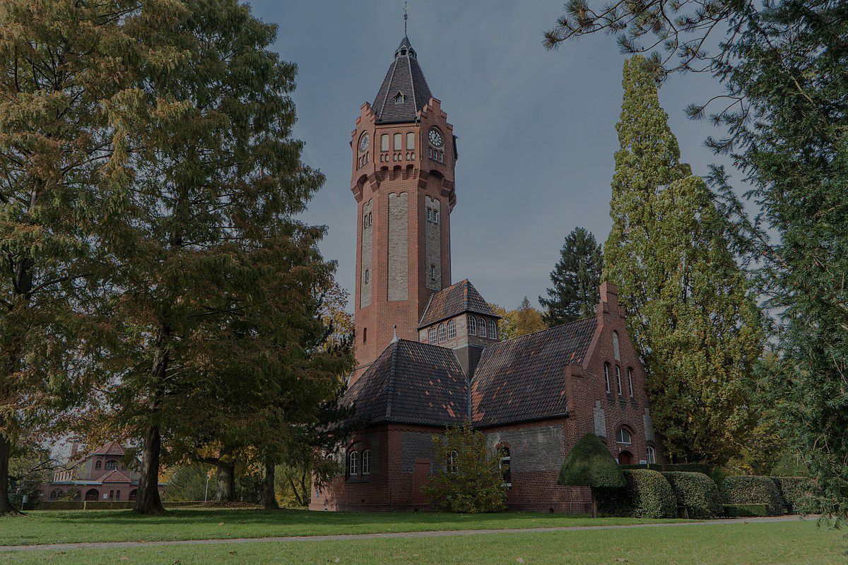 Bild des Wasserturms. Zwischen alten hohen Laubbäumen ist der Wasserturm aus Backstein zu sehen.