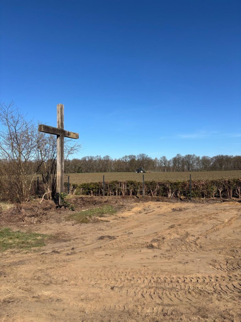 Blick über den Friedhof in Richtung eines Feldes. Links ist ein hohes Holzkreuz zu sehen. Die Fläche vor dem Kreuz ist sandiger Boden mit Spuren von Baggerreifen.