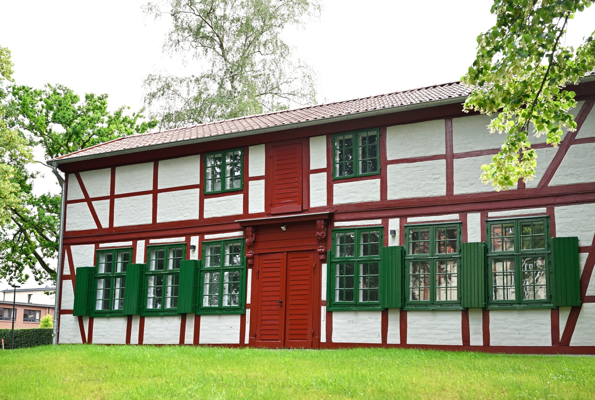 Es ist ein Foto des alten Gärtnerhauses. Es ist ein flaches zweistöckiges Fachwerkgebäude. Die Fensterläden sind dunkelgrün. Die Wände sind weiß und das Fachwerk dunkelrot.